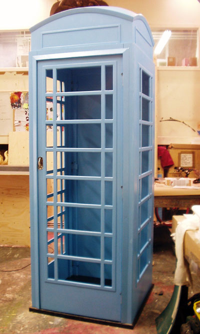 An old-style telephone box with glass doors. Painted in a primer coat of pale blue-grey